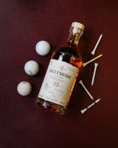 A bottle of Aultmore 25yo. Pictured on a maroon leather background with gold balls and tees.