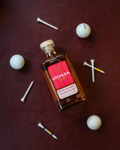 A bottle of Lochlea Harvest edition. Pictured on a maroon leather background with gold balls and tees.